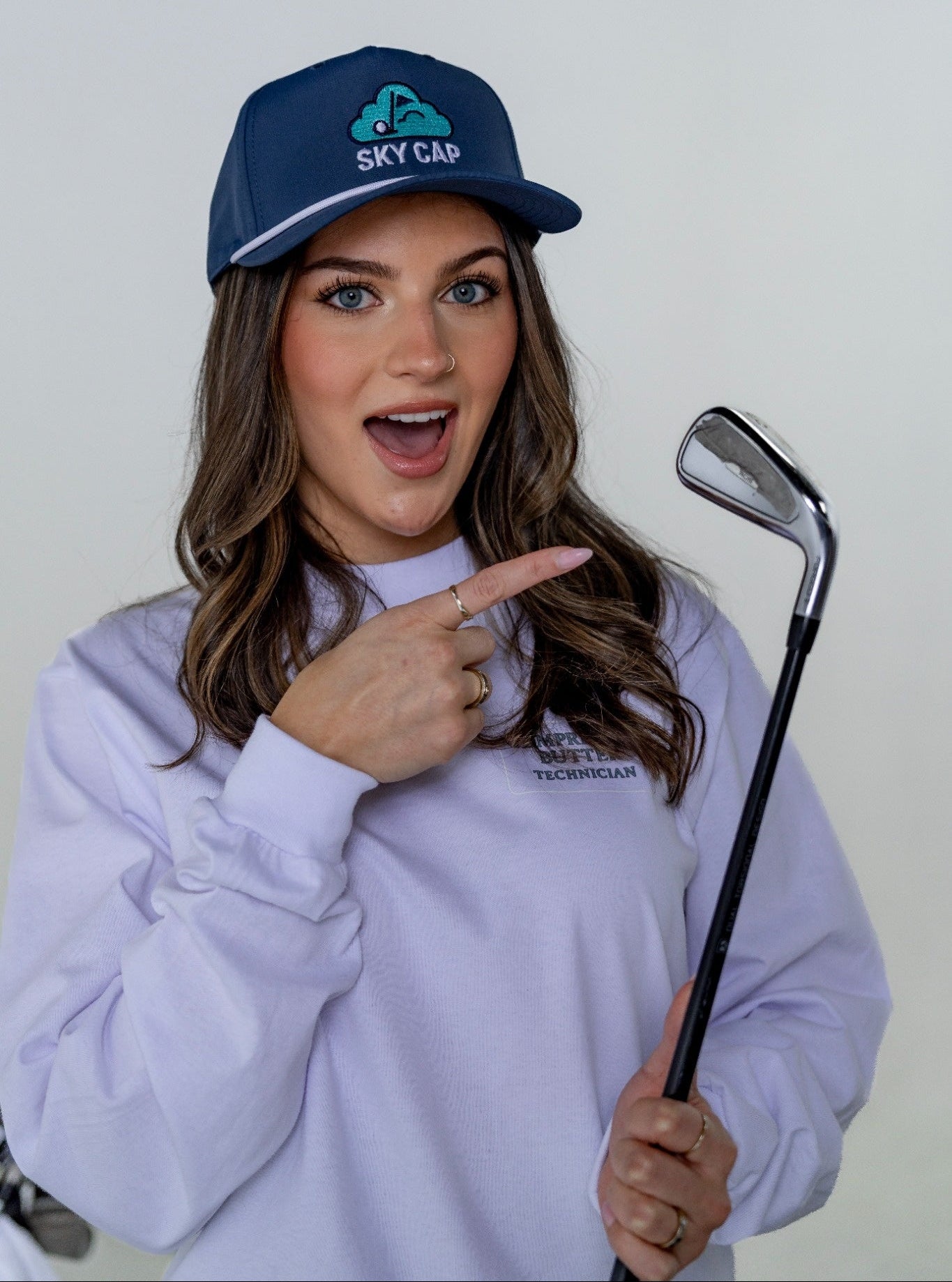 Woman wearing a blue cap and White Compressed Butter technician long sleeve, holding a golf club and wearing a Navy Sky Cap 