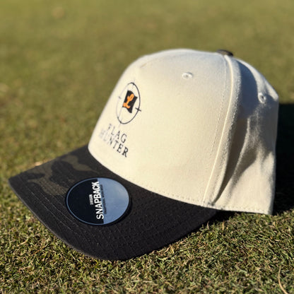 Beige and black Camo cap with a flag hunter logo on a grassy background