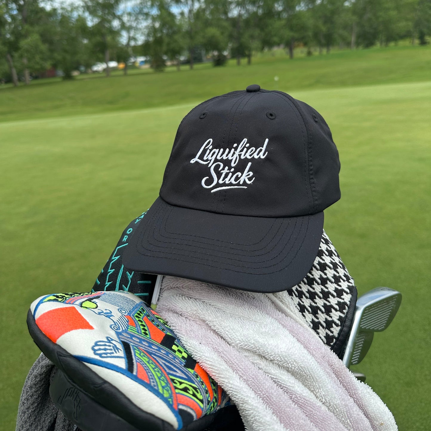 Black cap with 'Liquorished Sticks' logo on a golf bag with a golf club against a green golf course background.