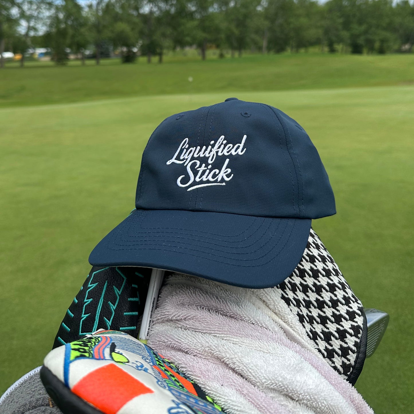 Navy cap with 'Liquified Stick' logo on a golf bag with green grass and blue sky in the background