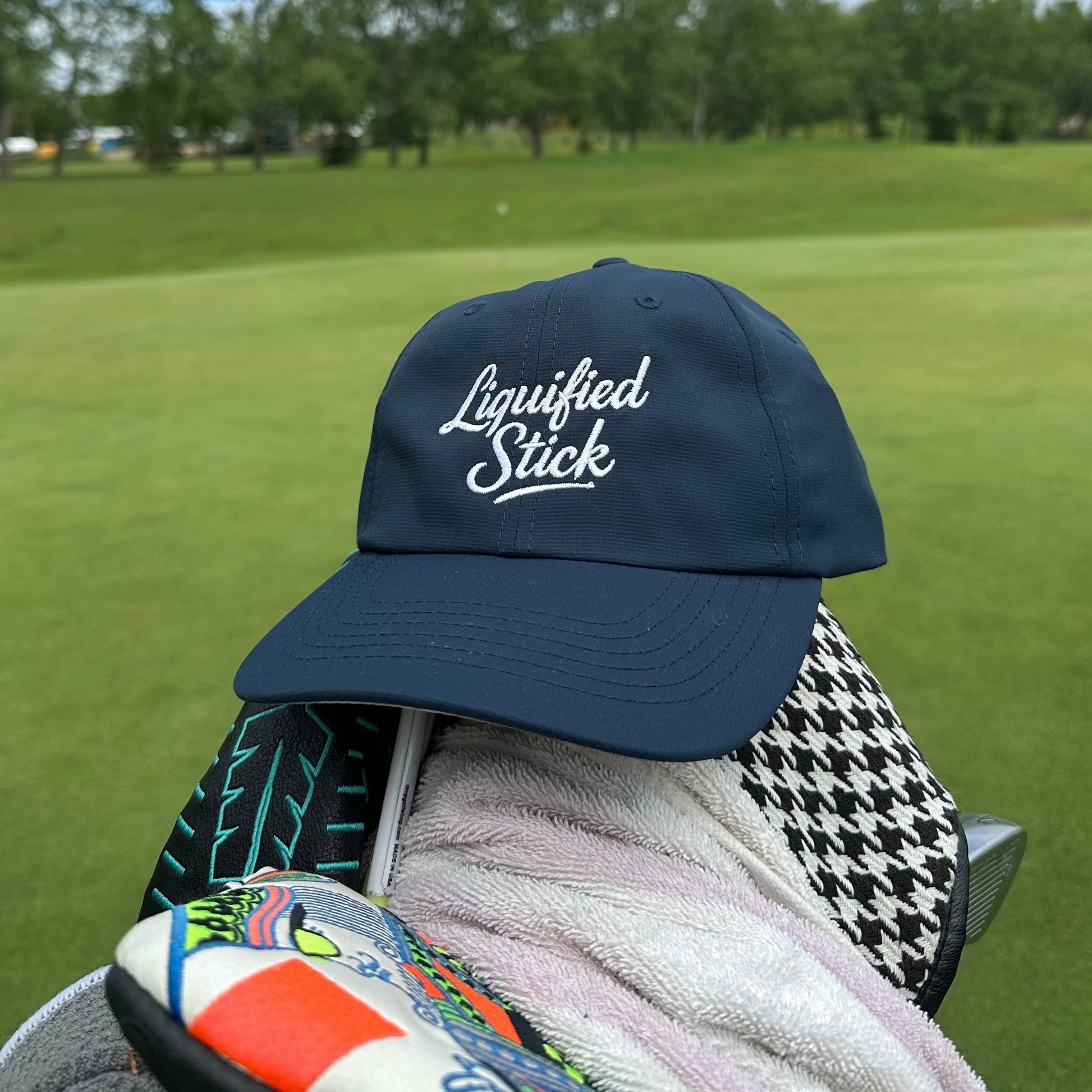 Navy cap with 'Liquified Stick' logo on a golf bag with green grass and blue sky in the background